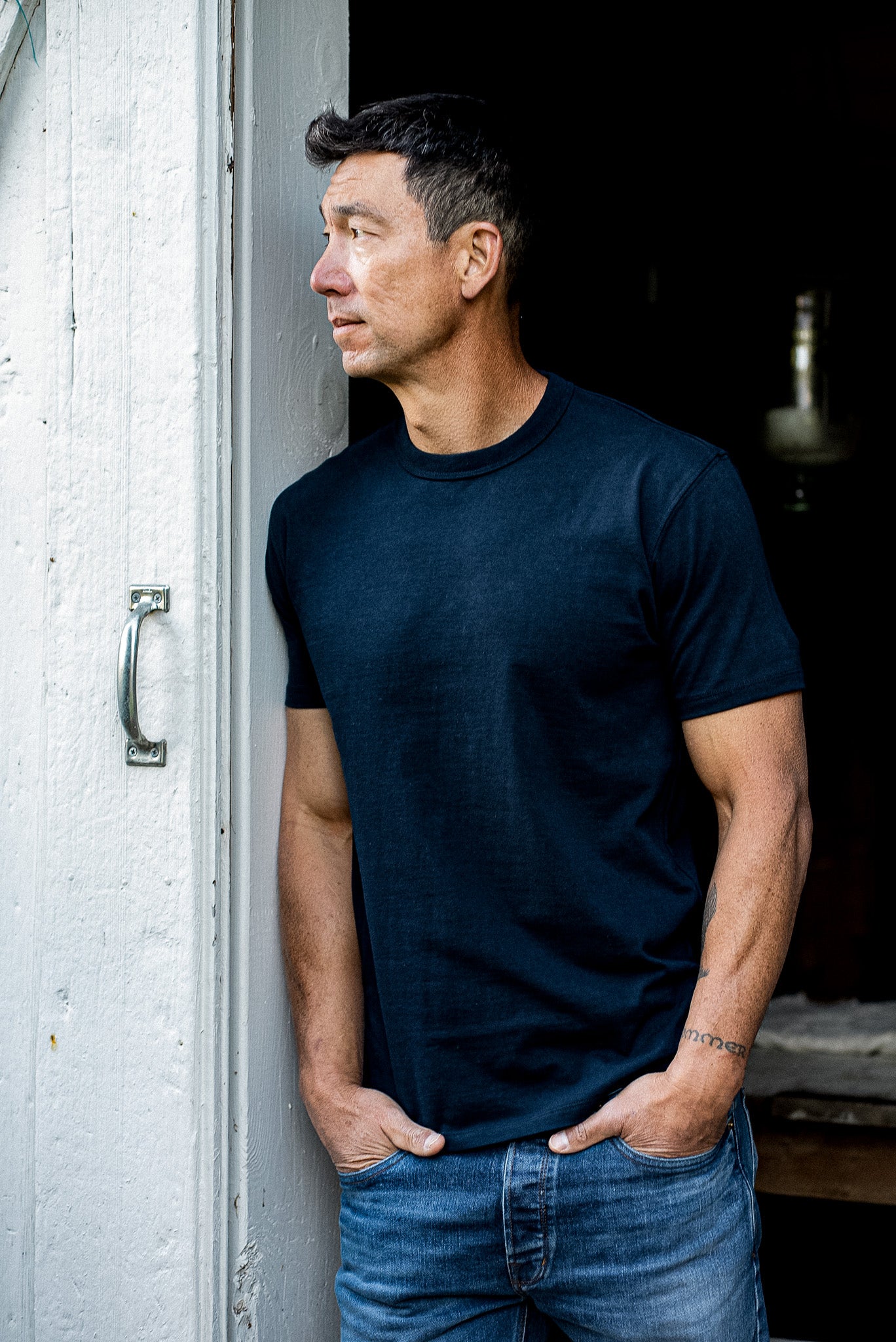 Man in a navy heavyweight cotton Bolinas tee and blue jeans leaning against a white doorway.