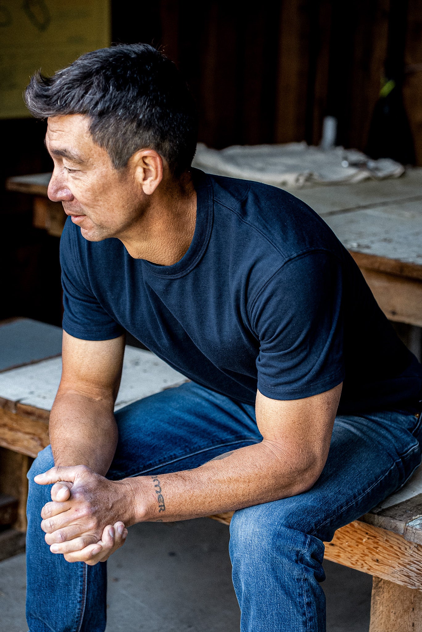 Man in a navy Bolinas heavyweight cotton tee and denim jeans seated on a wooden workbench.