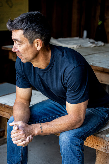 Man in a navy Bolinas heavyweight cotton tee and denim jeans seated on a wooden workbench.