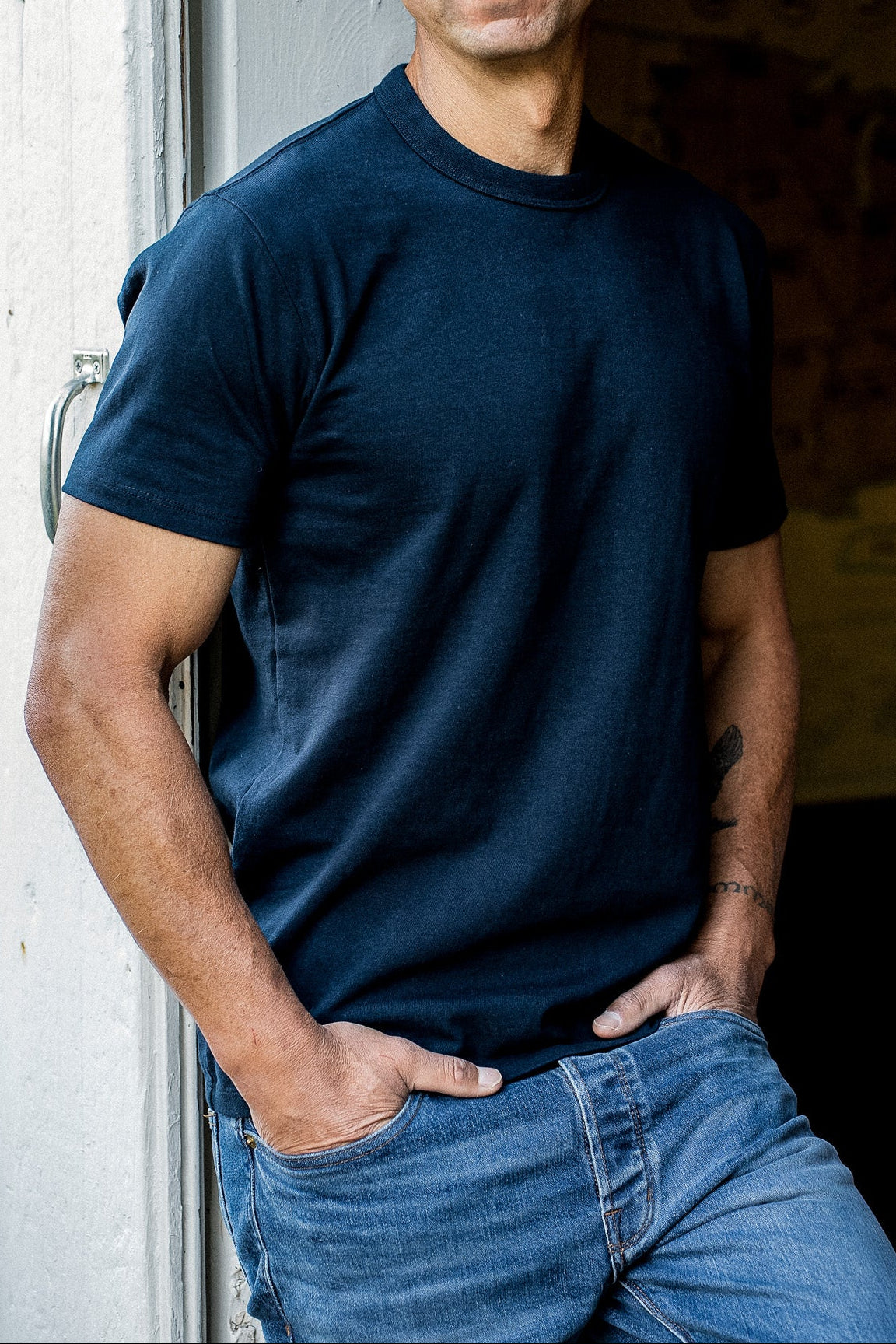 Man in navy heavyweight cotton Bolinas tee and blue jeans leaning against a weathered doorway.