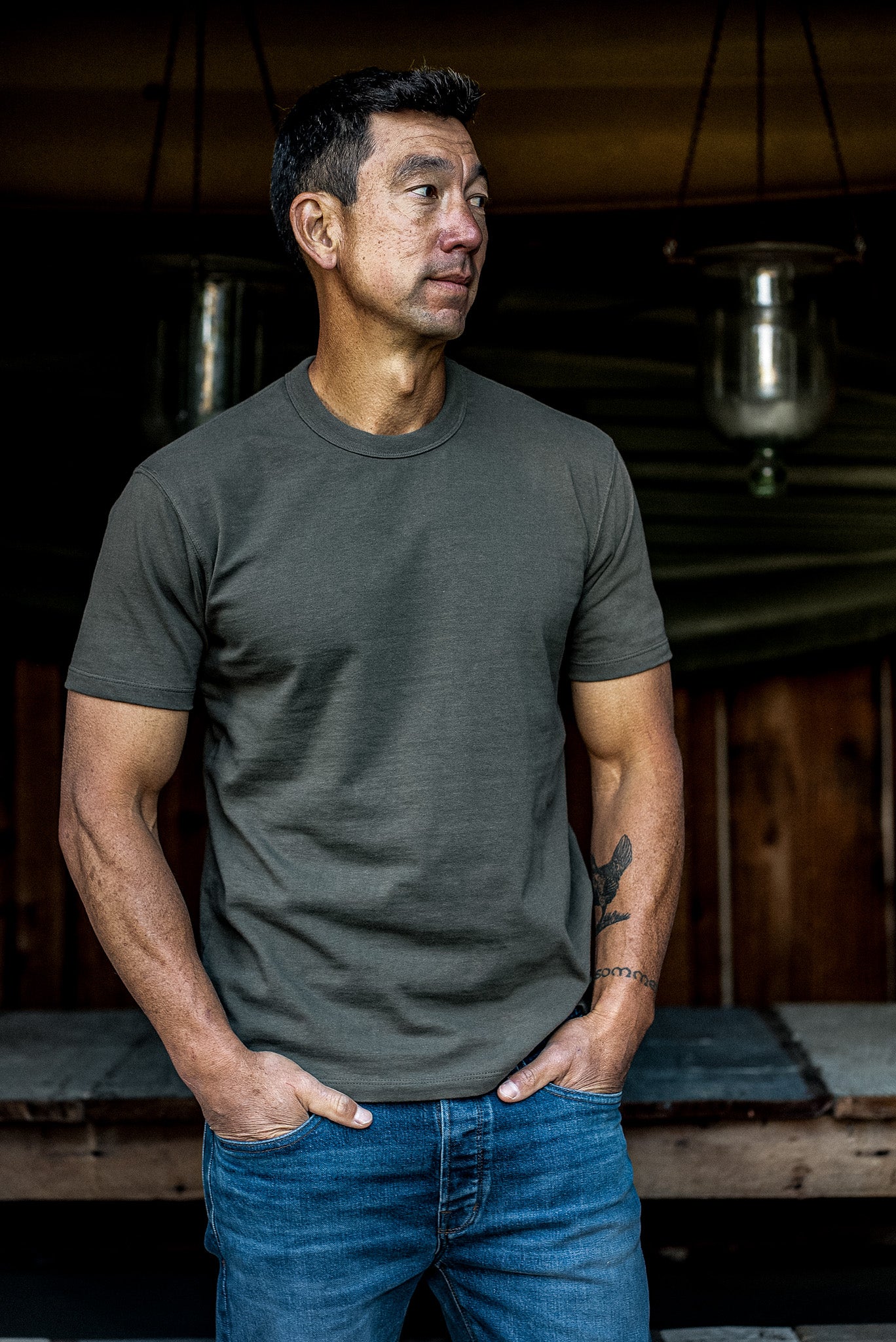 Man wearing the olive Bolinas heavyweight cotton tee with blue jeans, standing outside a wooden cabin.