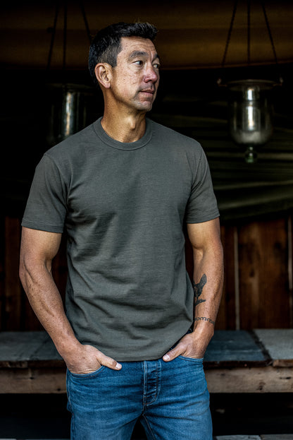 Man wearing the olive Bolinas heavyweight cotton tee with blue jeans, standing outside a wooden cabin.