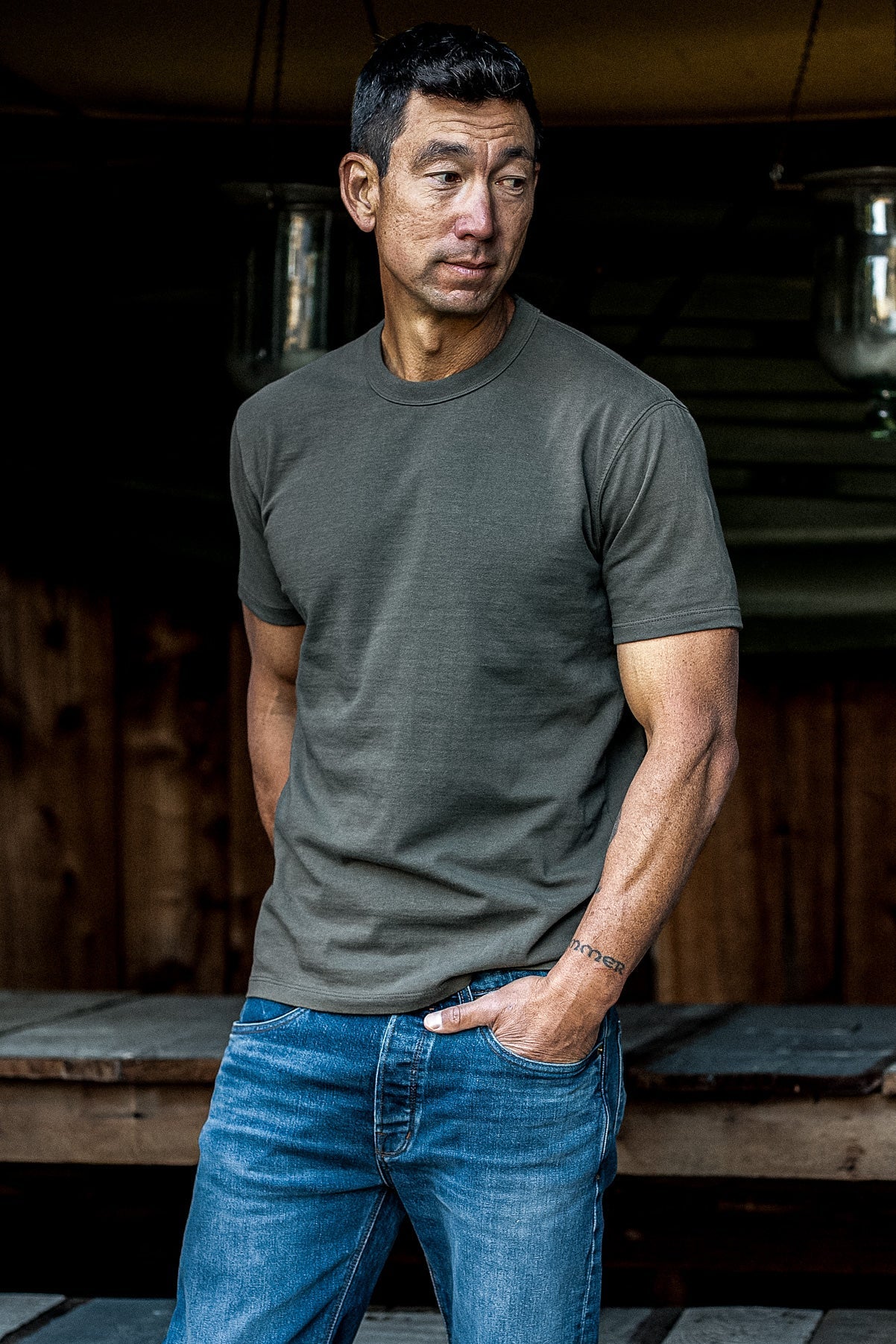 Man wearing an olive heavyweight Bolinas tee with blue jeans in front of a wooden cabin.