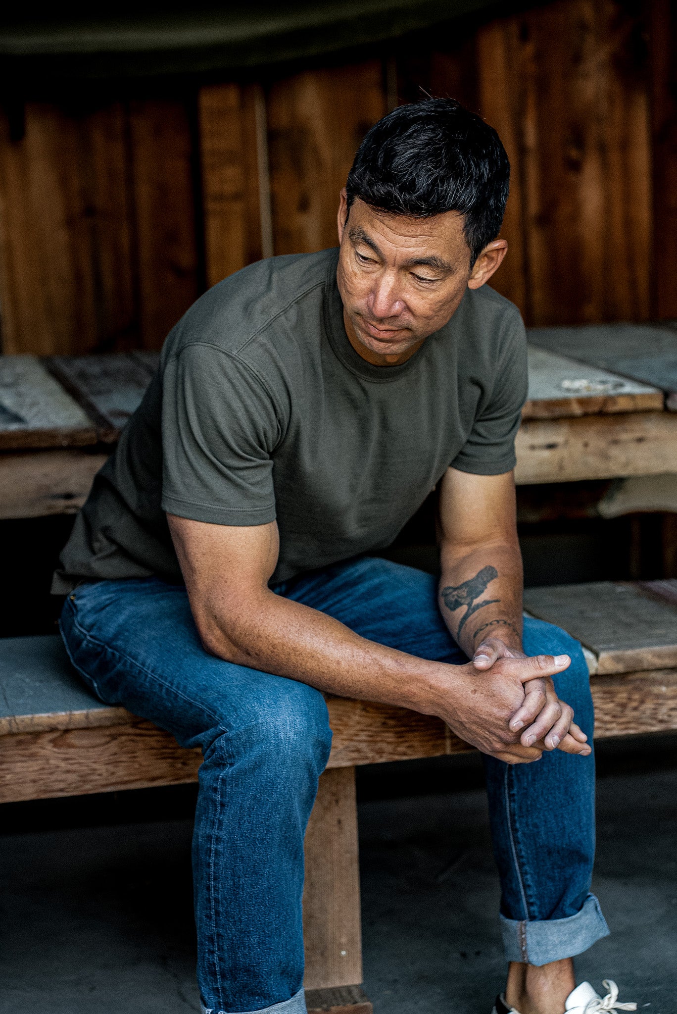 Man in olive heavyweight Bolinas tee and cuffed blue jeans seated on a wooden bench.