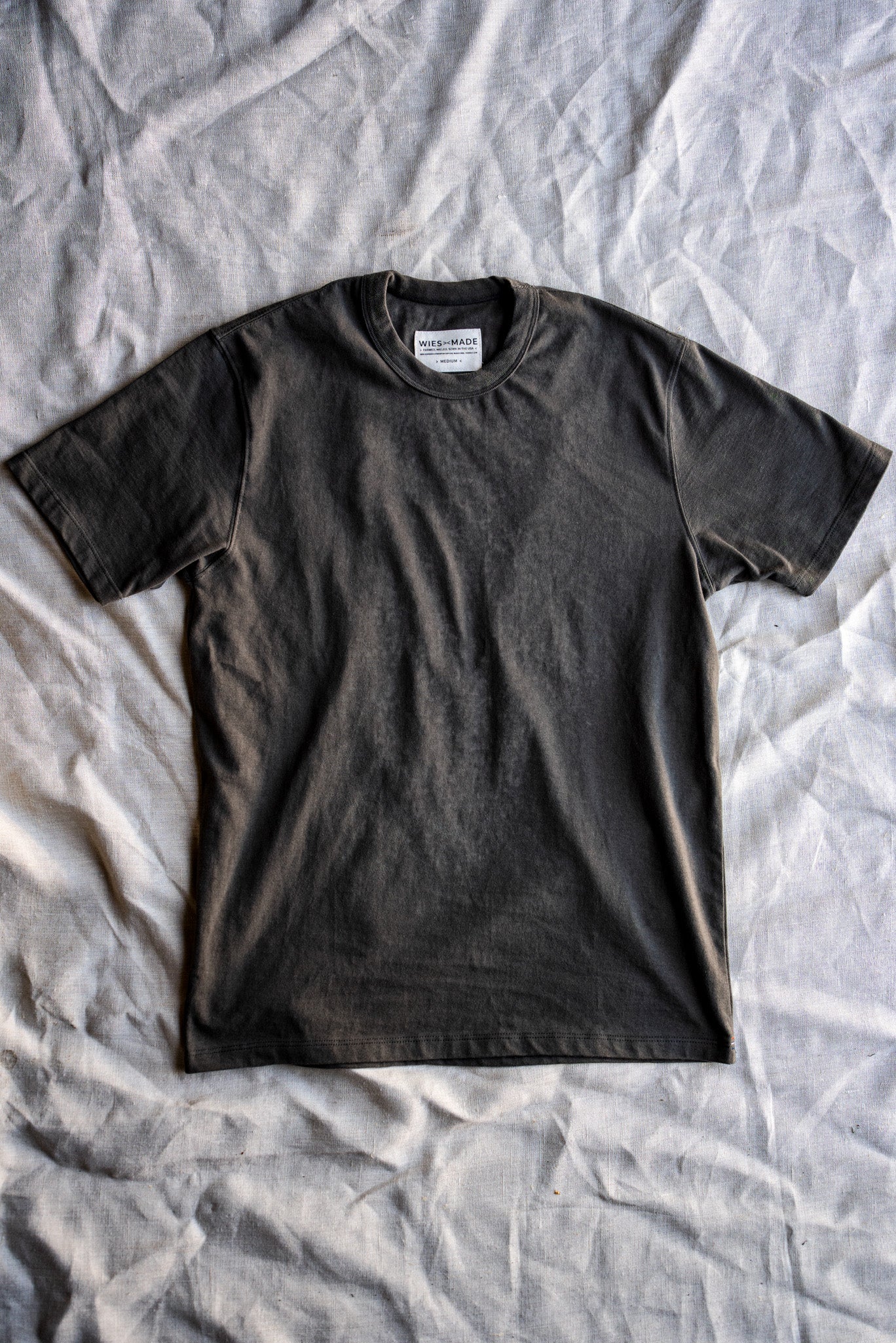 Olive heavyweight cotton Bolinas tee laid flat on a crumpled linen sheet, WiesMade neck label visible.