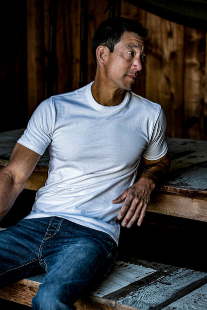 Man in white heavyweight cotton Bolinas tee and jeans, seated against rustic wood planks.