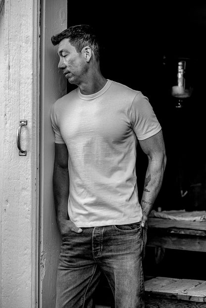 Man in a natural cotton Bolinas heavyweight tee and jeans leaning against a weathered doorway.