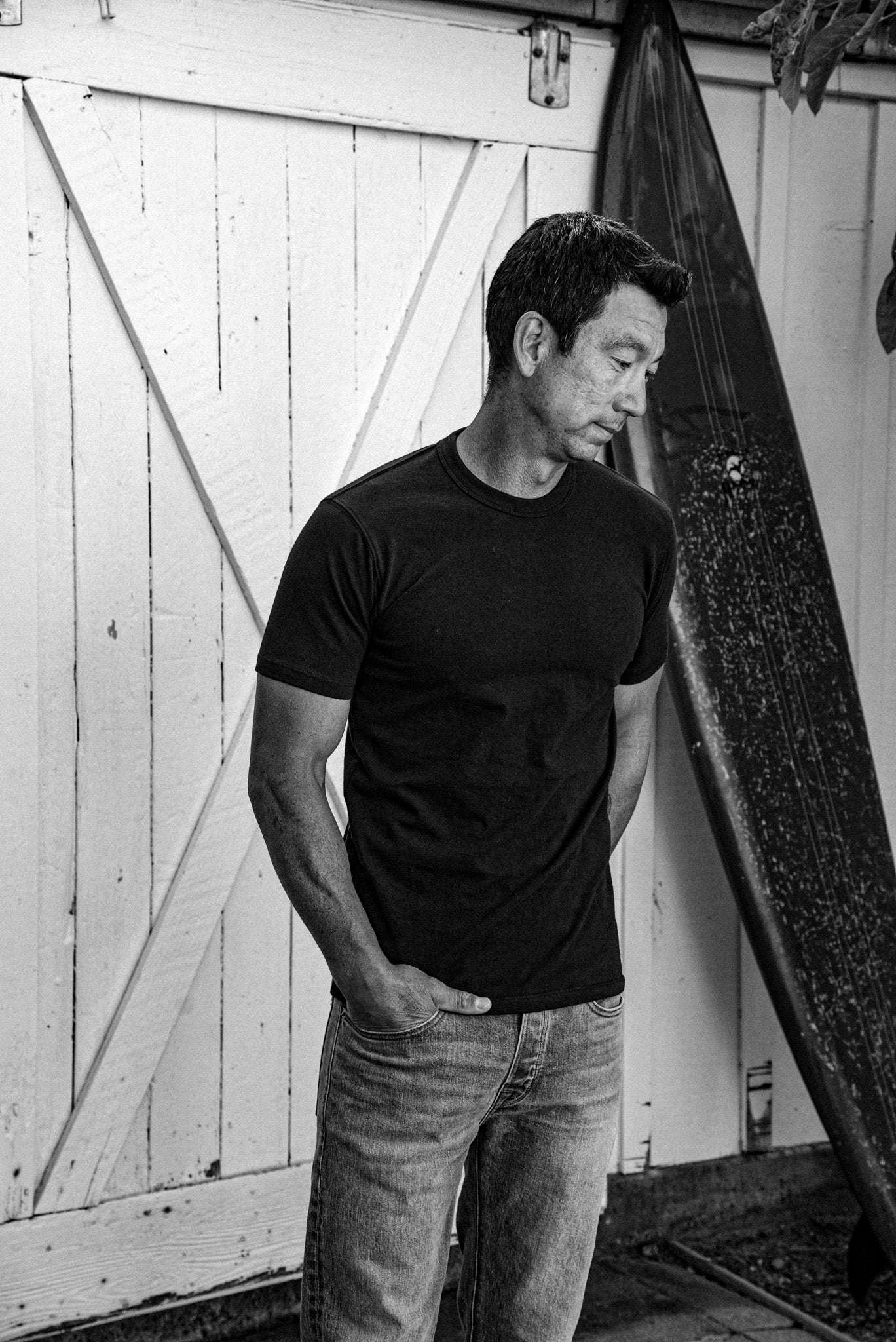 Man in a Bolinas heavyweight tee and jeans beside a surfboard and weathered barn door.