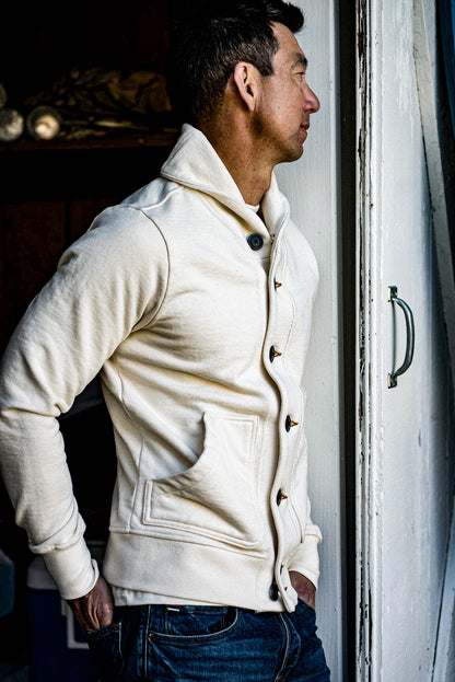 Side view of natural cream shawl collar French terry cardigan worn with denim jeans in a doorway.