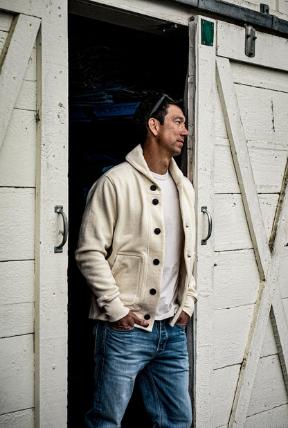 Man in natural cream shawl collar French terry cardigan and blue jeans leaning in a white barn doorway.