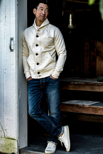 Man leaning in a doorway wearing a natural cream shawl collar French terry cardigan with selvedge denim jeans.
