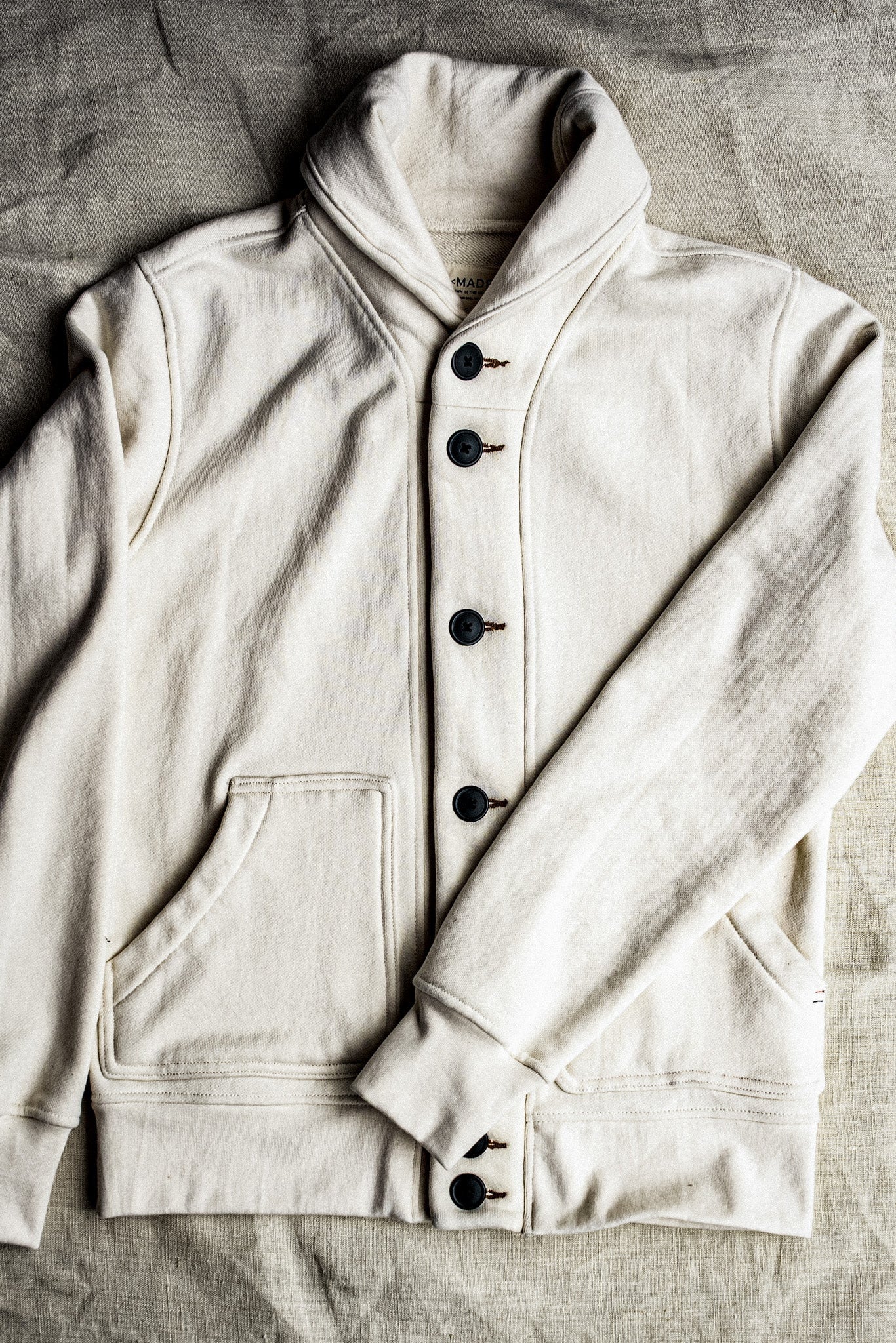 Natural cream shawl collar cardigan in cotton French terry with dark buttons, laid flat on linen.