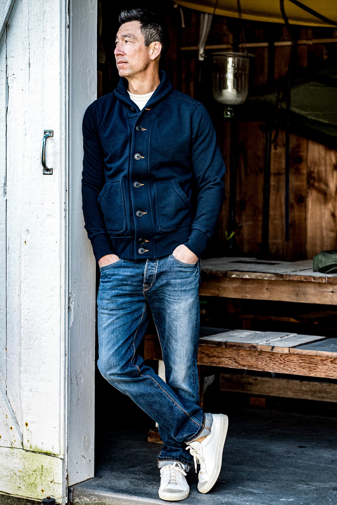 Man leaning in a doorway wearing a navy shawl-collar French terry cardigan with selvedge denim jeans.