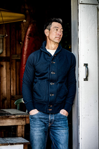 Man in a navy shawl collar Bodega cardigan and blue jeans leaning in a workshop doorway.