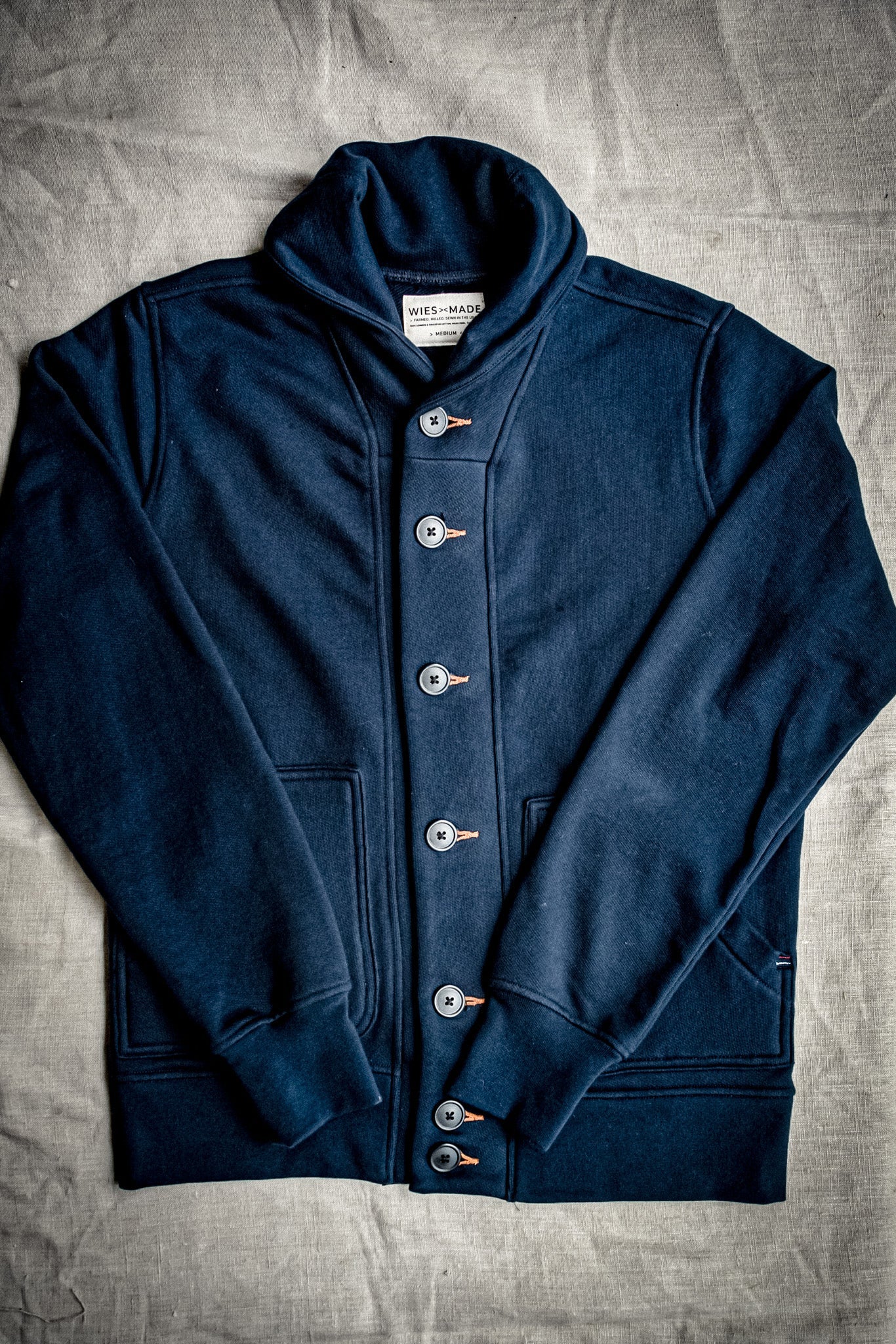 Navy French terry shawl-collar cardigan laid flat, with tobacco-thread buttonholes and front pockets.