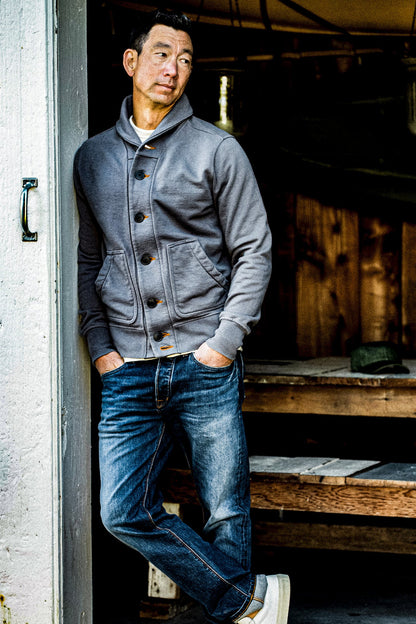 Man leaning in a doorway wearing a pebble grey shawl collar French terry cardigan with selvedge denim jeans.