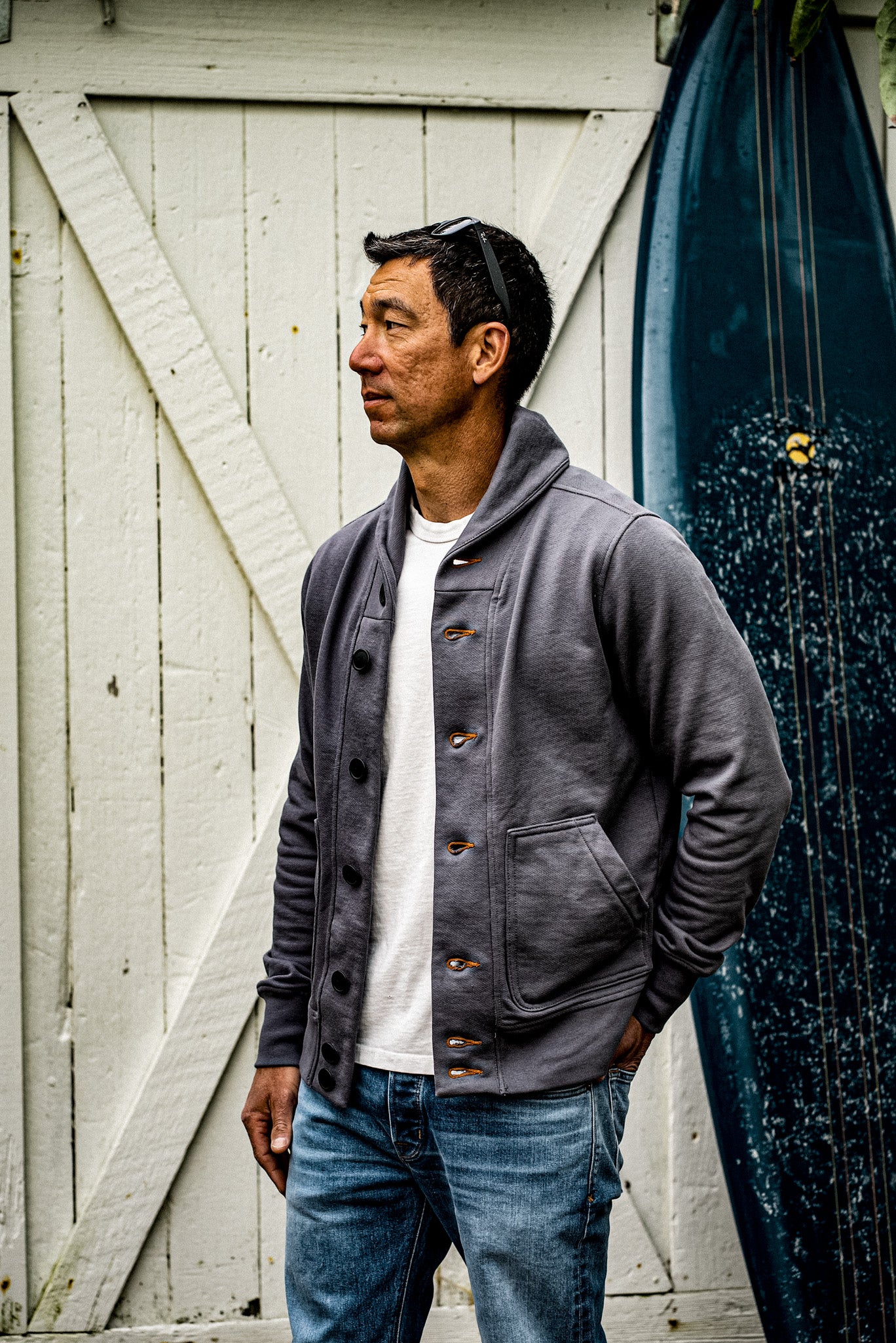 Man in a pebble grey shawl collar French terry cardigan with tobacco keyhole buttonholes, worn over a white tee and jeans by.