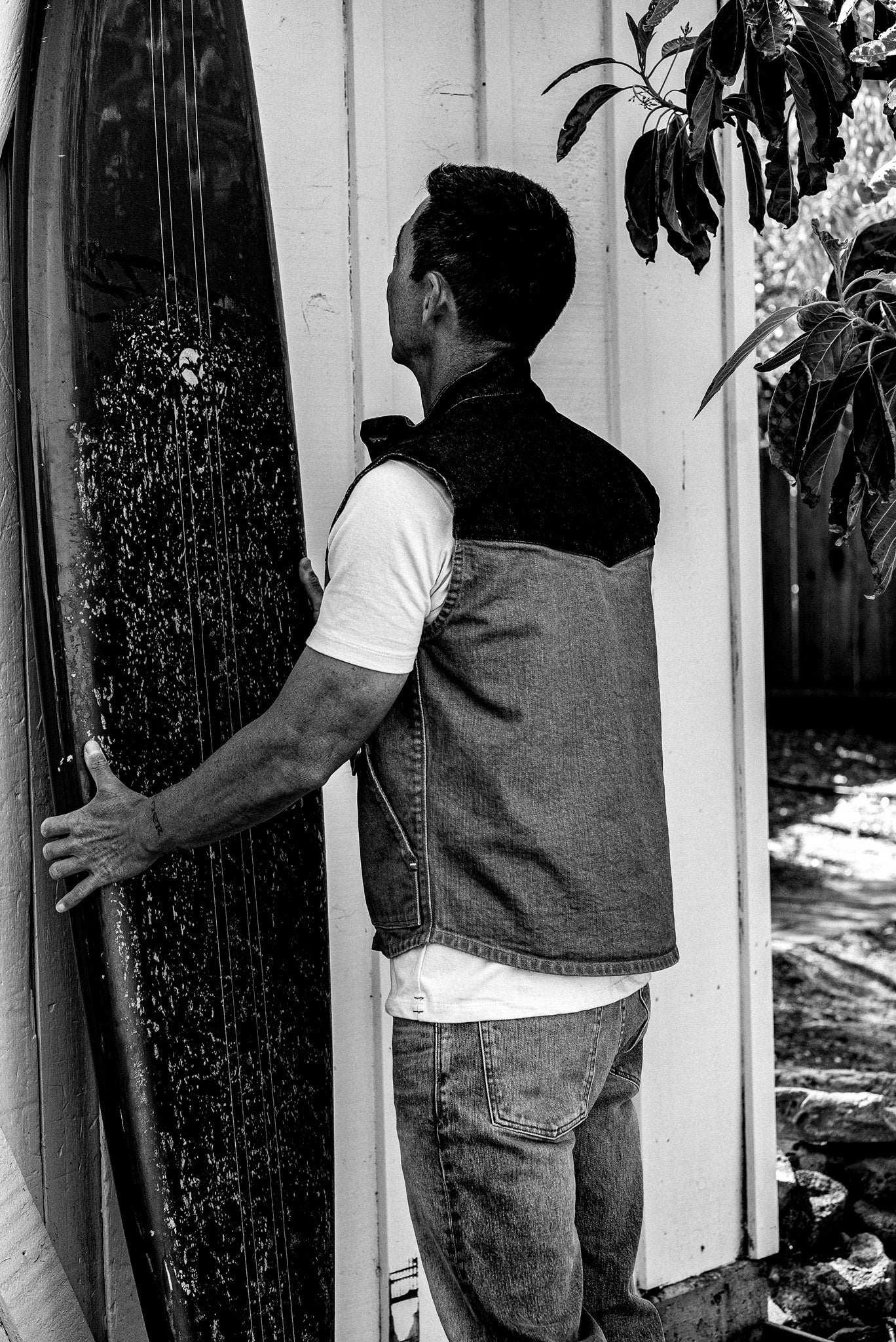 Man in a two-tone denim vest and jeans holding a longboard surfboard beside a wooden shed.