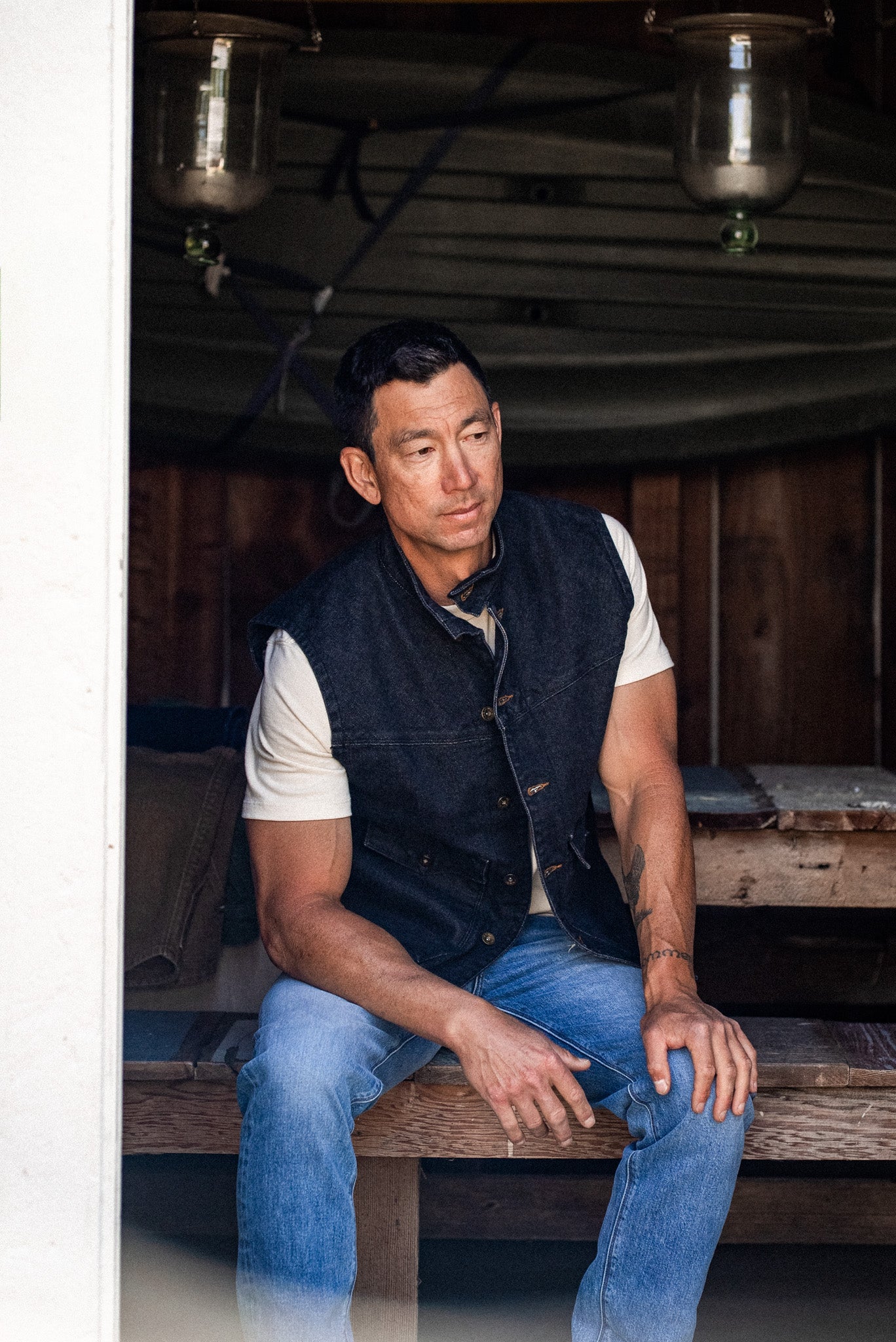 Man in black broken twill denim vest and blue jeans sitting on a workbench in a wood-paneled shed.