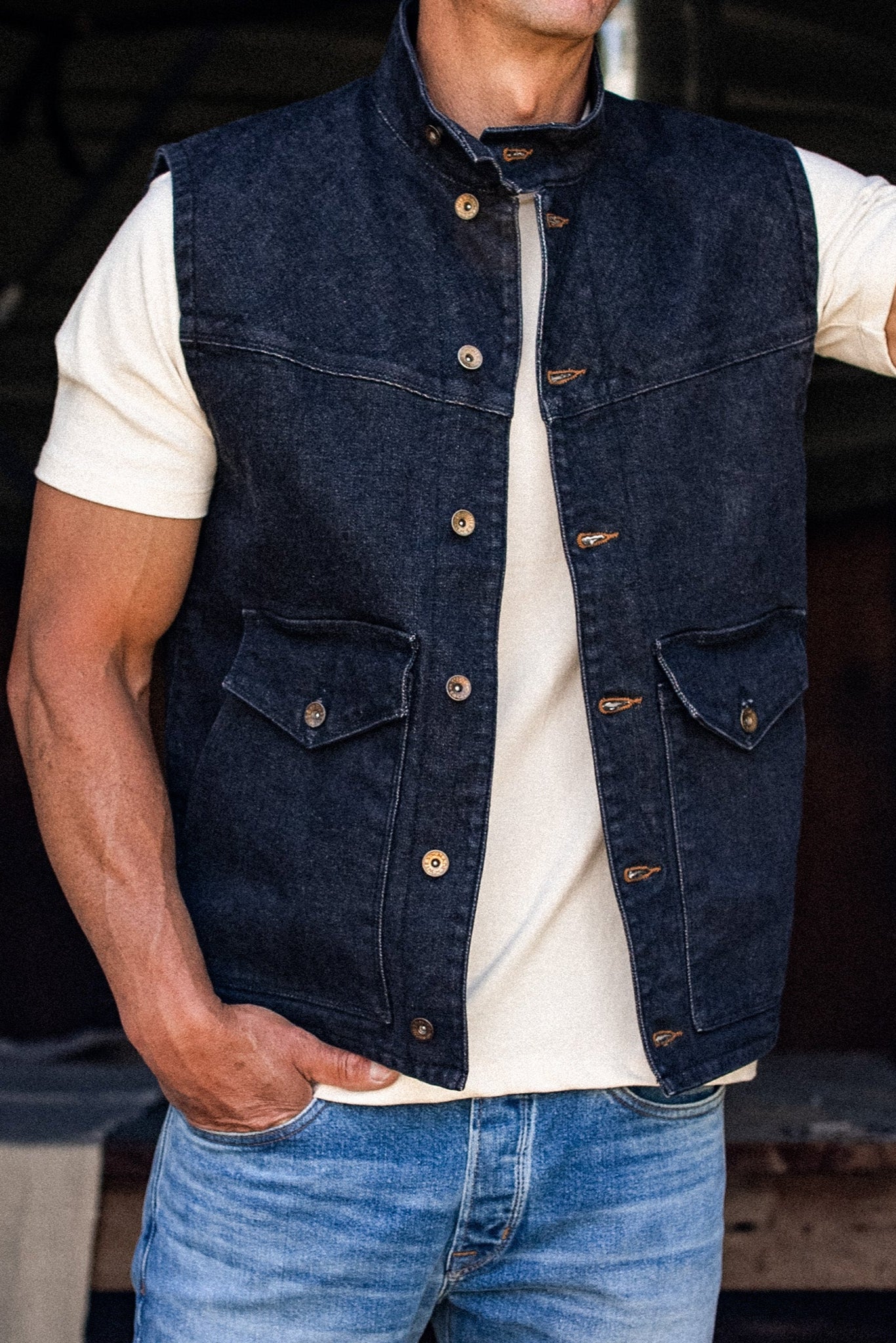 Black broken twill denim vest worn open over a cream tee with blue jeans, showing brass snaps and button-flap chest pockets.