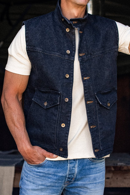 Black broken twill denim vest worn open over a cream tee with blue jeans, showing brass snaps and button-flap chest pockets.