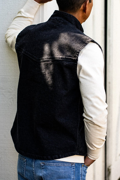 Back view of a black broken twill denim vest worn over a cream long-sleeve shirt with blue jeans.