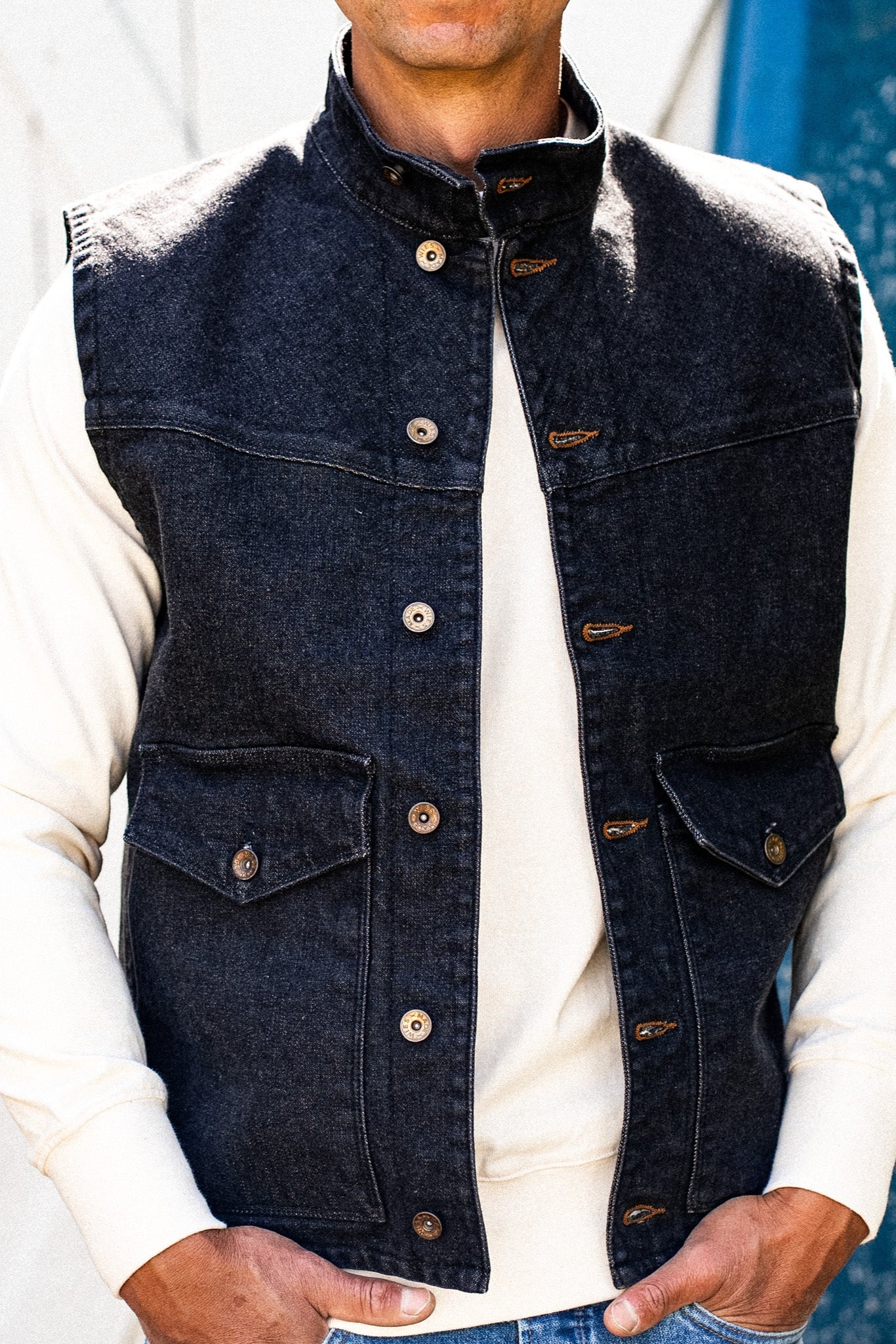 Black 14oz broken twill denim vest with stand collar, brass buttons, and flap pockets worn over a cream sweatshirt.