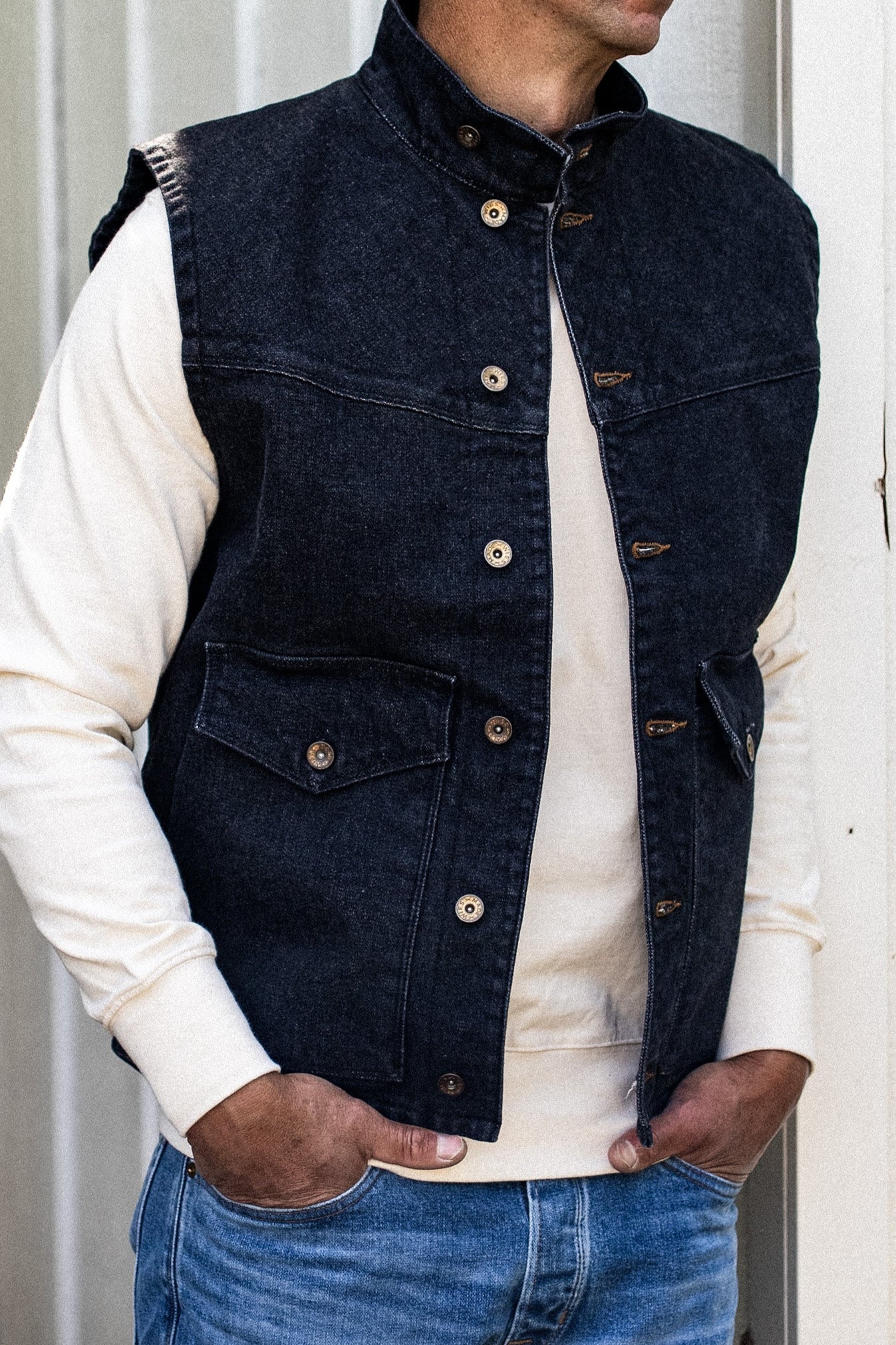 Black broken twill denim vest with stand collar and brass snaps, worn over a cream sweatshirt and blue jeans.