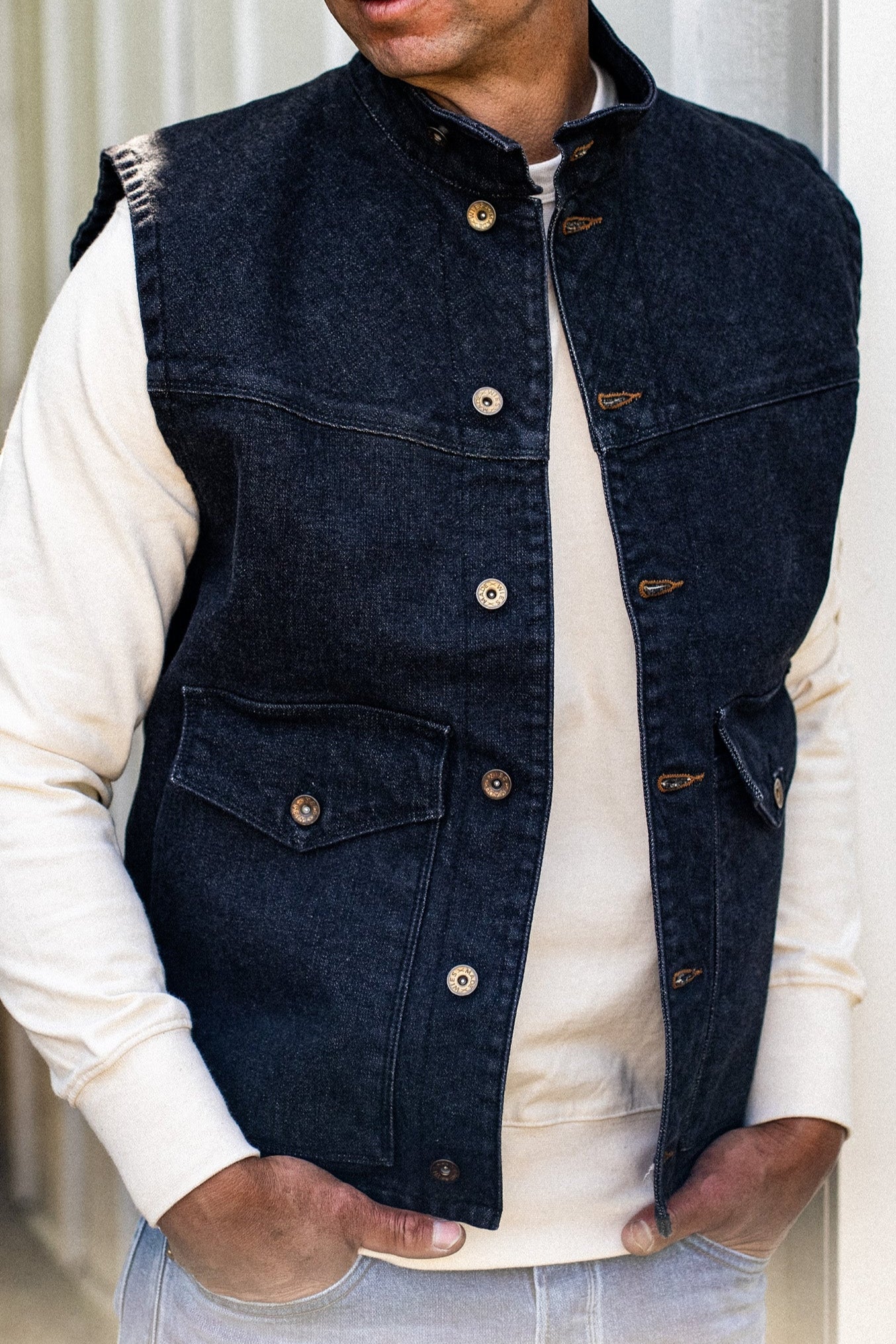 Black broken twill denim vest worn open over a cream sweatshirt, showing brass buttons and flap chest pockets.