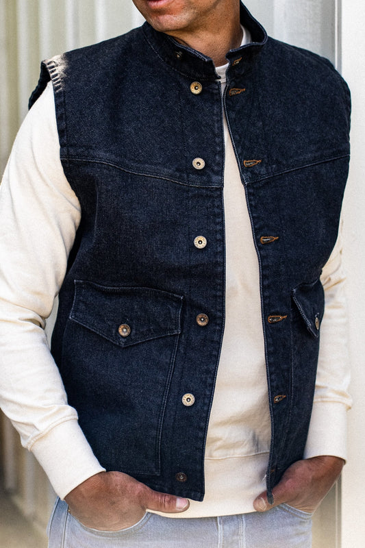 Black broken twill denim vest worn open over a cream sweatshirt, showing brass buttons and flap chest pockets.