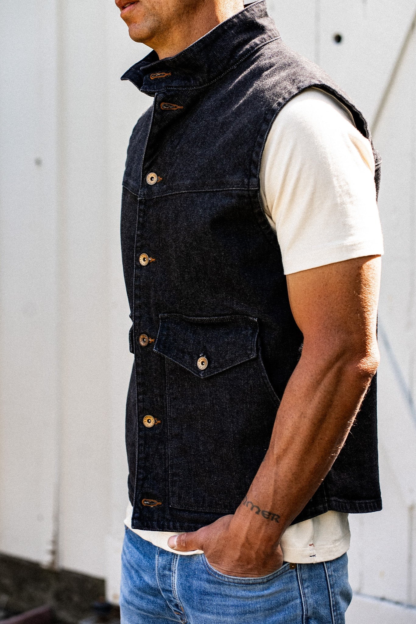 Side view of model wearing the black broken twill denim vest over a cream tee with blue jeans.