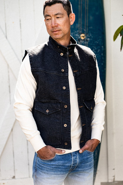 Man wearing black broken twill denim James Vest over cream sweater outdoors.