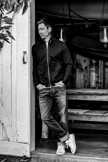Man in a black cotton Stinson moto full-zip jacket, leaning against a weathered doorway.