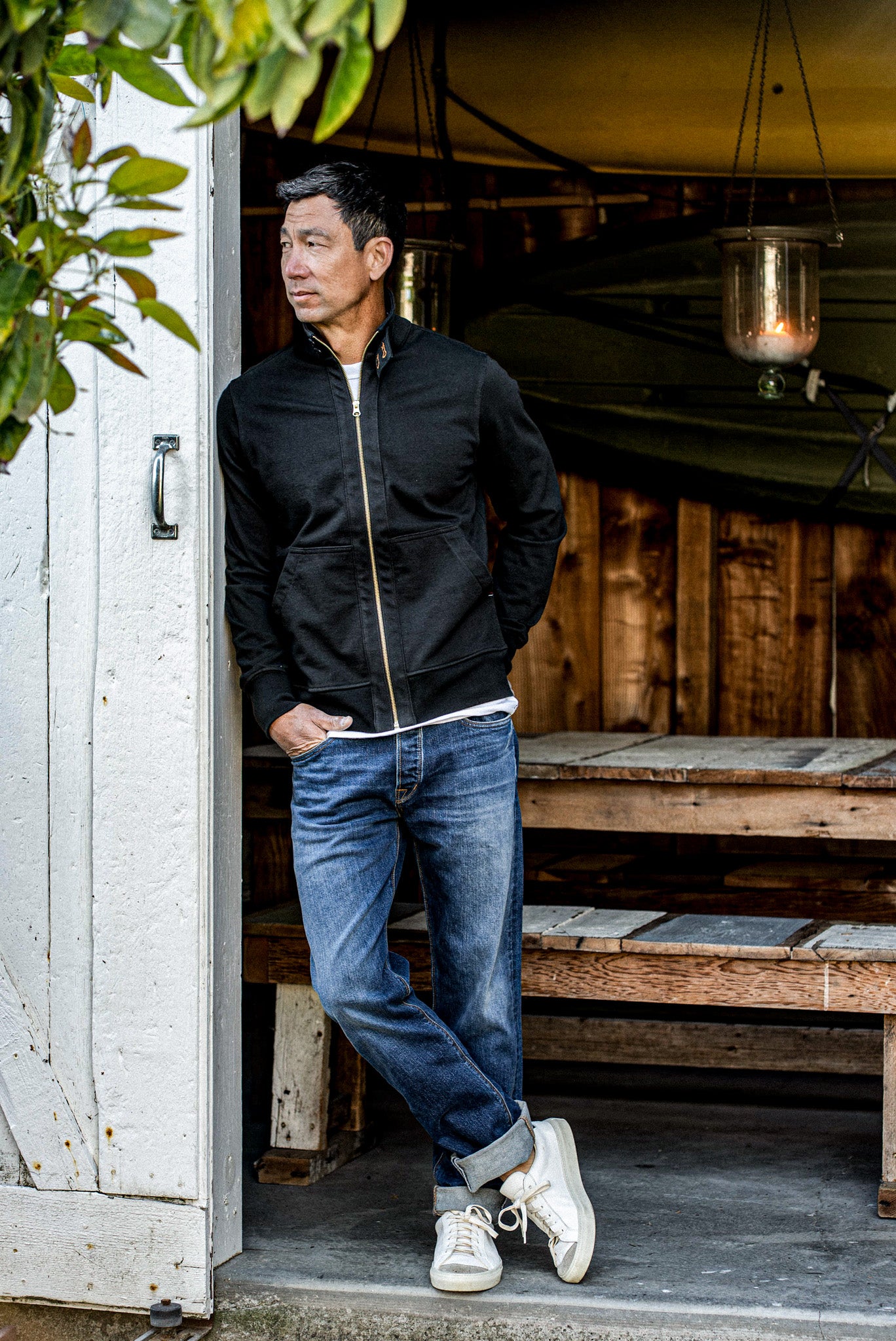 Black cotton moto jacket with brass zipper, worn open over a tee and jeans in a doorway.