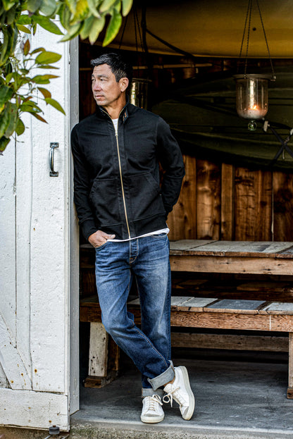 Black cotton moto jacket with brass zipper, worn open over a tee and jeans in a doorway.