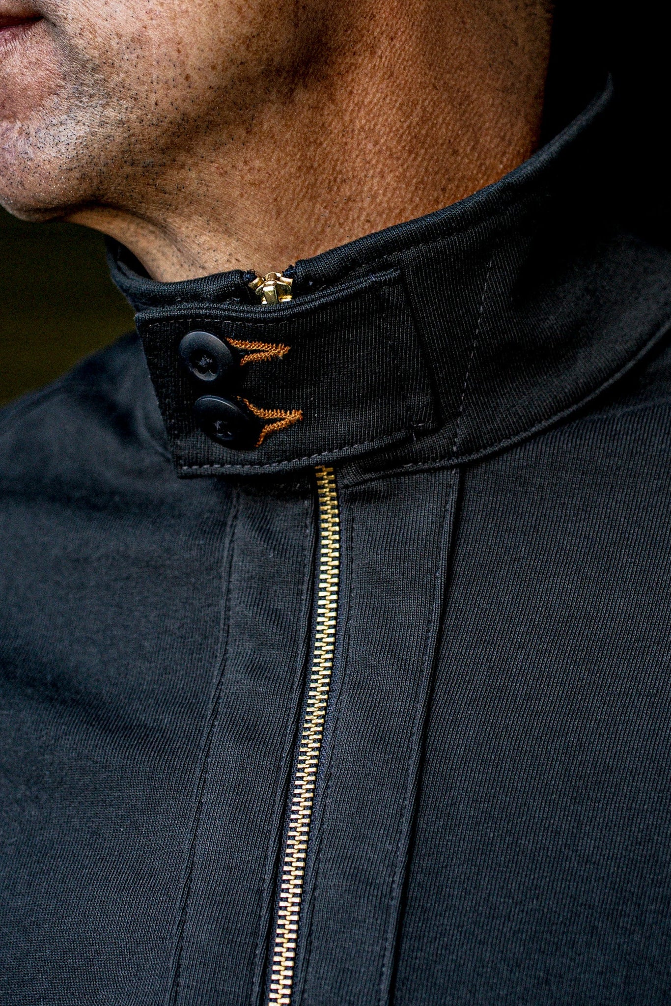 Stand collar detail of basalt cotton moto jacket with brass zipper and orange contrast stitching.