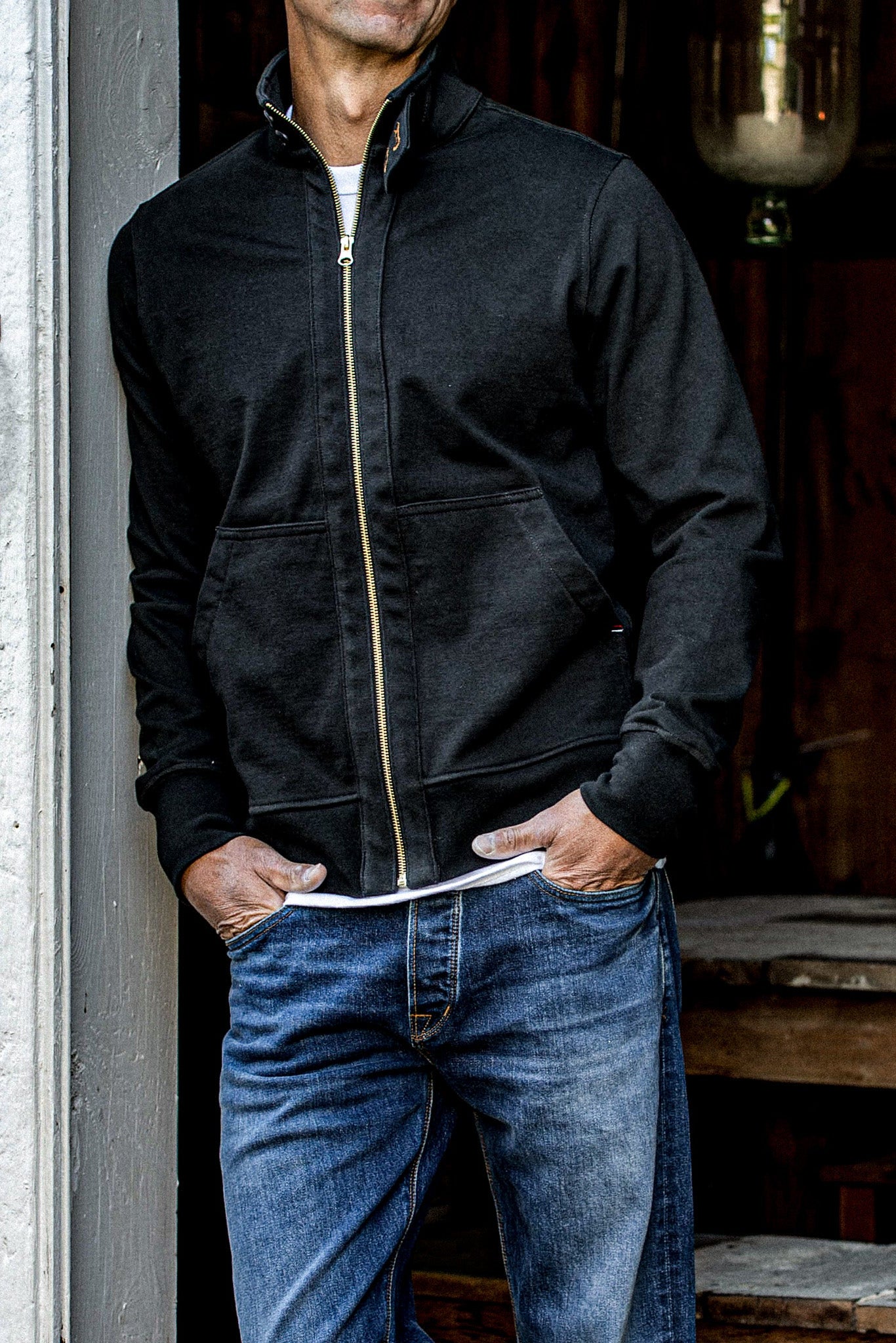 Man wearing a basalt cotton moto jacket with brass front zipper, hands in jean pockets.
