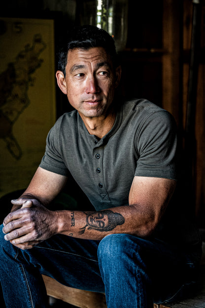 Man in an olive men's Bolinas Henley shirt with four-button placket, seated indoors.