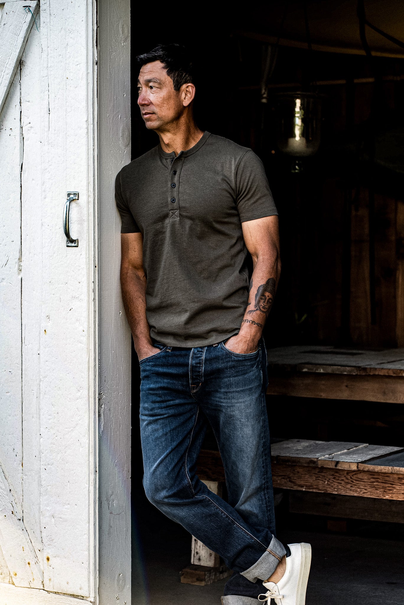 Man in olive short-sleeve Bolinas Henley and cuffed denim jeans leaning in a weathered doorway.