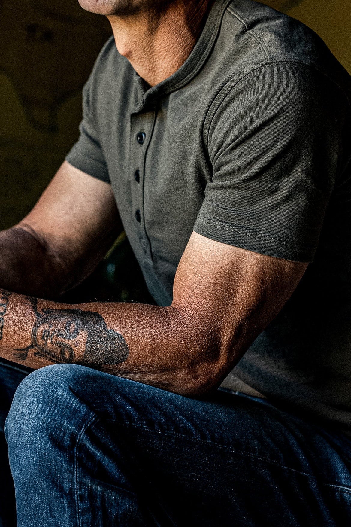 Man seated in an olive four-button Bolinas Henley paired with dark denim jeans.