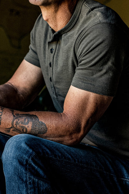 Man seated in an olive four-button Bolinas Henley paired with dark denim jeans.