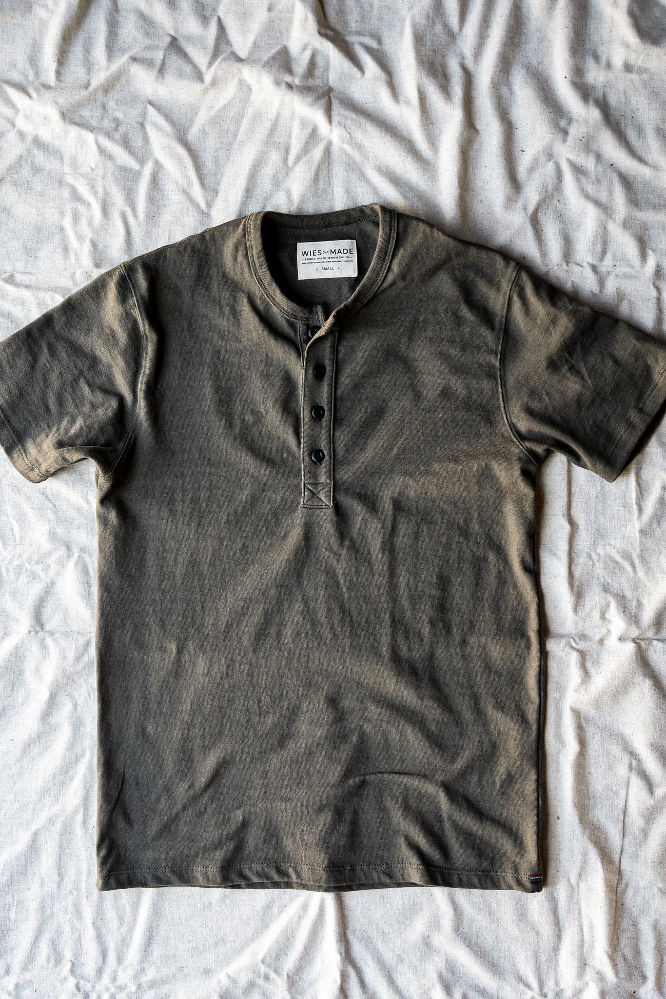 Olive green short-sleeve Bolinas Henley with four dark buttons, laid flat on a white cotton sheet.