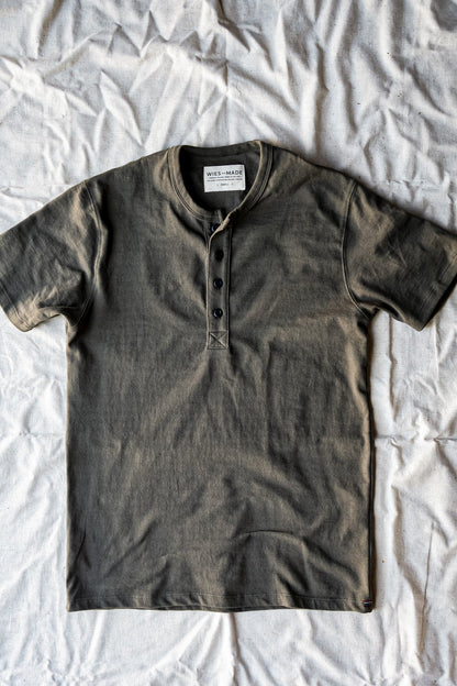 Olive green short-sleeve Bolinas Henley with four dark buttons, laid flat on a white cotton sheet.