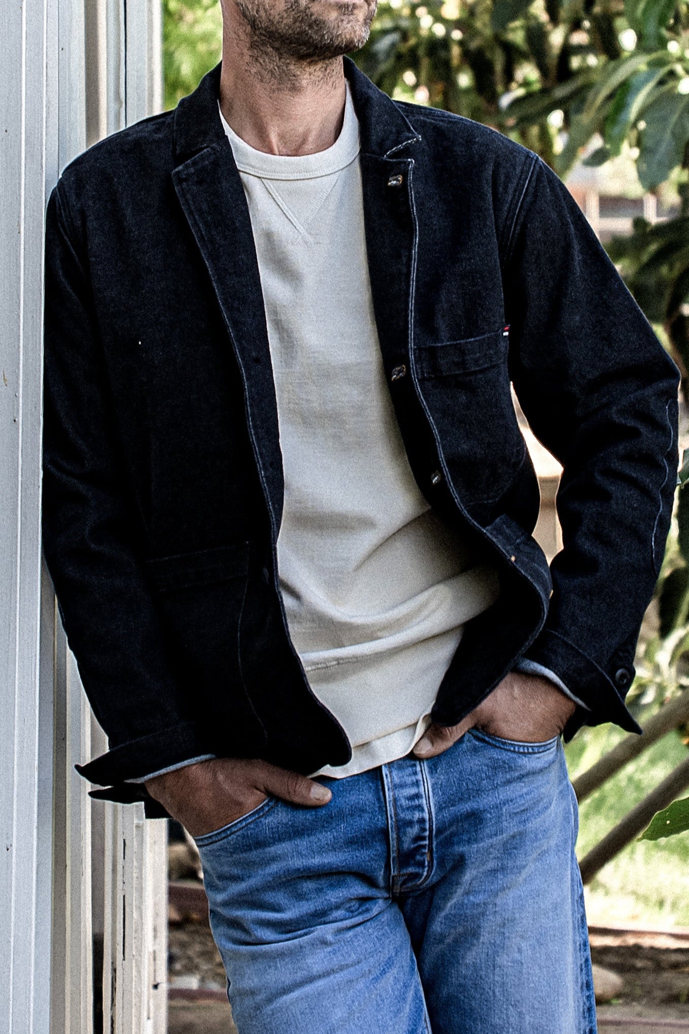 Man in a black denim chore blazer worn open over a cream sweatshirt and blue jeans.