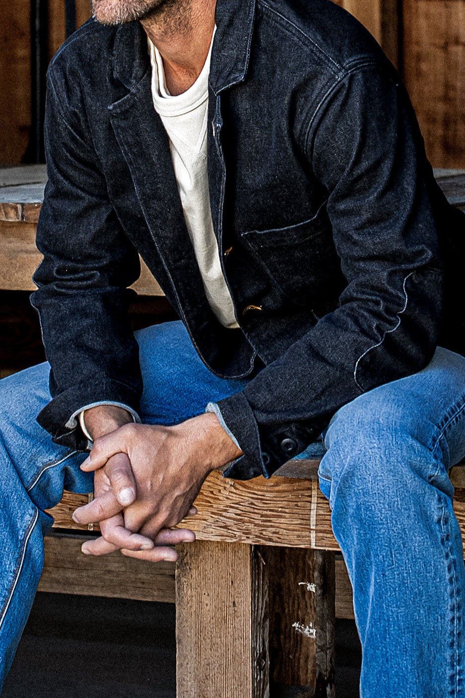 Man in a black denim chore blazer and blue jeans seated on a wooden bench.