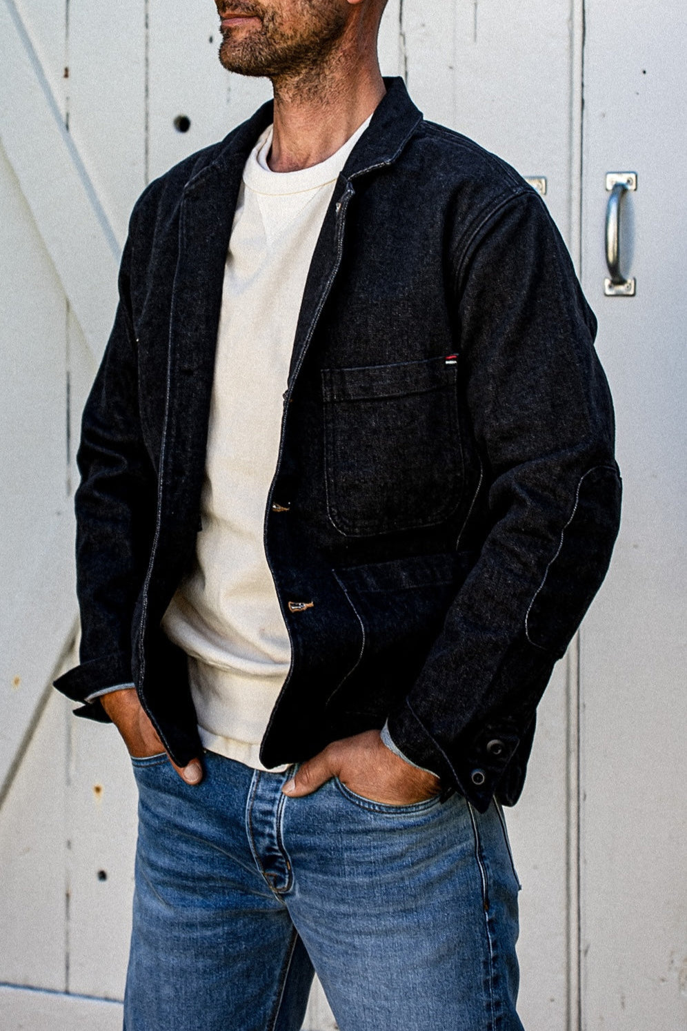 Man wearing a black denim chore blazer open over a cream tee with light-wash jeans.