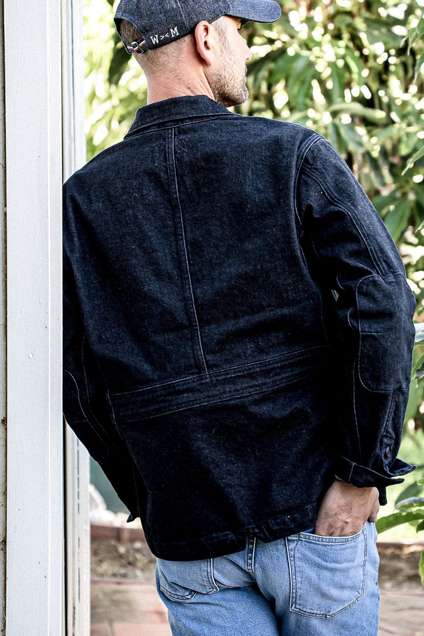 Back view of a man wearing a black denim chore blazer with elbow patches, paired with light blue jeans.