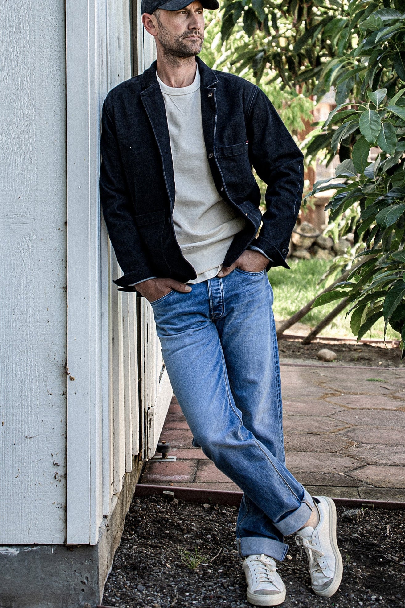Man in black Scout denim chore blazer over tee, leaning against a weathered doorway outdoors.