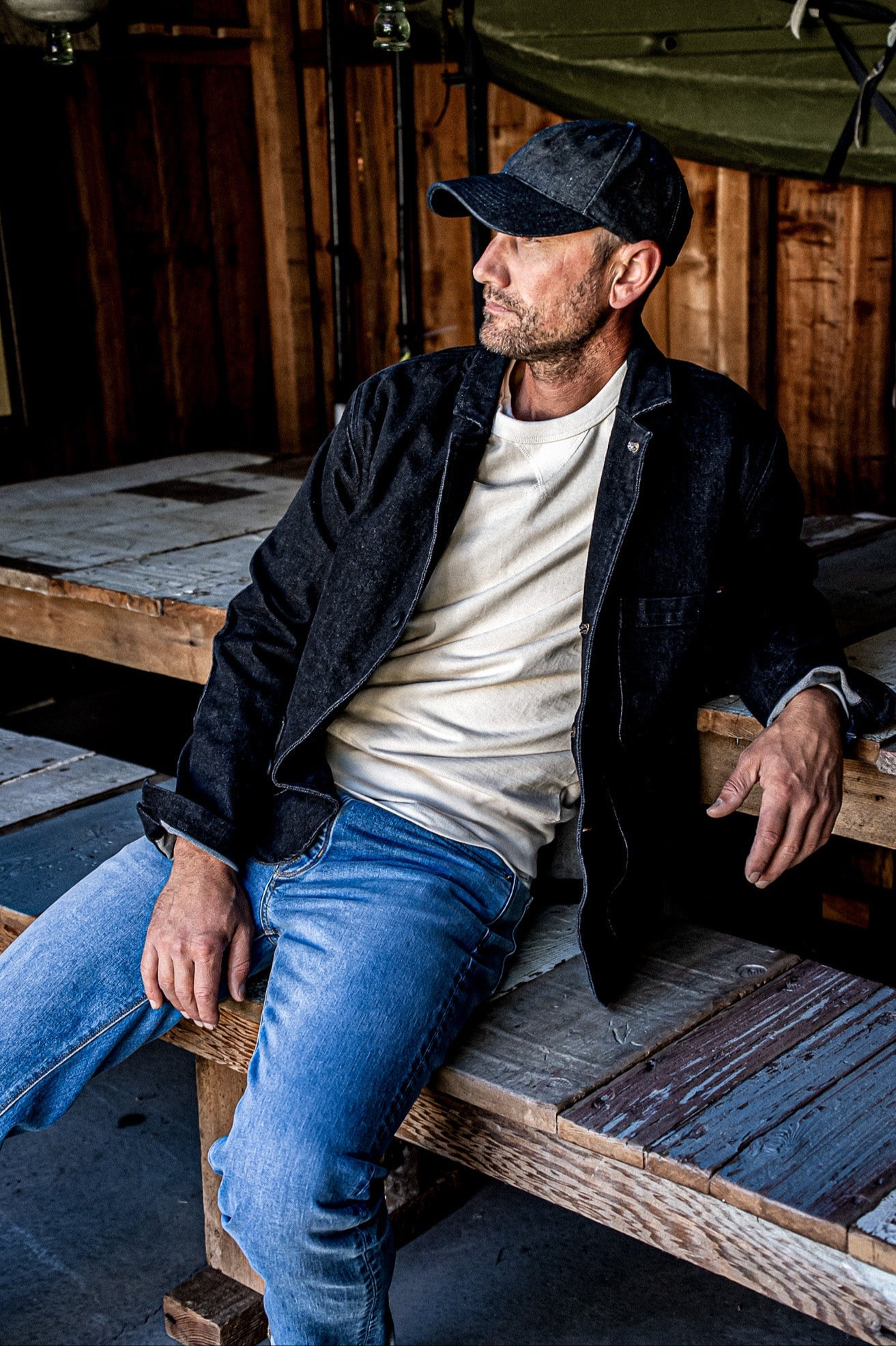 Man in a black denim chore blazer over a cream tee and blue jeans, seated on a wooden pallet in a rustic barn.