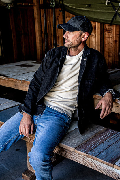 Man in a black denim chore blazer over a cream tee and blue jeans, seated on a wooden pallet in a rustic barn.